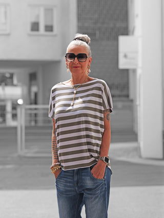 Ältere Frau trägt das OFF#DLY Marion Stripe Tee white/khaki, ein locker geschnittenes gestreiftes Kurzarmshirt, kombiniert mit blauer Jeans und stilvollen Accessoires, in urbaner Kulisse.