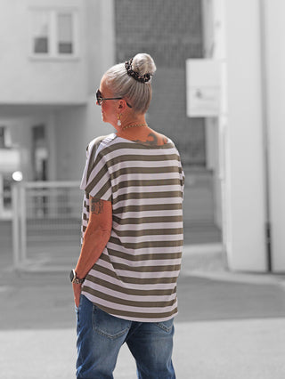 Ältere Frau trägt OFF#DLY Marion Stripe Tee weiß/khaki mit breiten Streifen, kurzen Ärmeln und lockerer Passform, kombiniert mit Jeans. Sichtbar sind goldene Accessoires und eine Smartwatch.