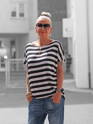 Frau in urbaner Umgebung trägt das OFF#DLY Marion Stripe Tee white/black, ein locker geschnittenes, schwarz-weiß gestreiftes T-Shirt, kombiniert mit blauer Jeans und goldfarbenem Schmuck.