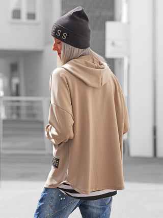 Person von hinten mit OFF#DLY Sweatjacke Hope taupe, locker geschnitten mit Kapuze unten, kombiniert mit schwarzer Beanie und blauen Jeans, in urbaner Umgebung. Ideal für lässigen Alltagslook.