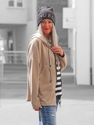Erwachsene Frau trägt die OFF#DLY Sweatjacke Hope taupe, eine hellbraune Kapuzenjacke mit Reißverschluss und Kordeln, über einem gestreiften Oberteil, kombiniert mit leicht zerrissenen blauen Jeans.