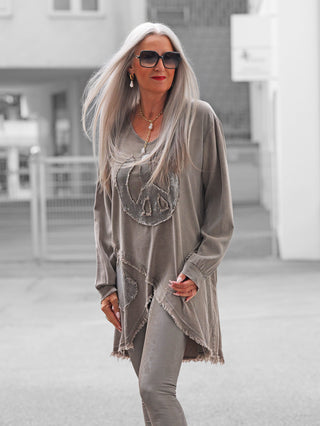 Frau in urbaner Umgebung trägt die OFF#DLY Tunica PEACE taupe mit großem glitzerndem Peace-Symbol, asymmetrischen Fransen und lässigem Schnitt, kombiniert mit schmalen, texturierten Leggings.