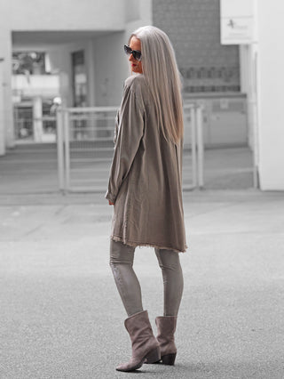 Person trägt den OFF#DLY Tunica PEACE in Taupe, einen lässigen, lang geschnittenen Mantel mit Fransen, über Leggings und Stiefeln, kombiniert mit Sonnenbrille für einen urbanen Vintage-Look.