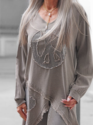 Die OFF#DLY Tunica PEACE taupe zeigt ein asymmetrisches Design mit rohen Kanten und funkelndem Pailletten-Patchwork, getragen mit einer dezenten Goldkette und passenden Leggings.