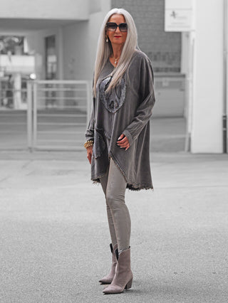 Reife Frau in OFF#DLY Tunica PEACE anthrazit mit fransigem Saum, Pailletten-Peace-Zeichen, übertaupe Leggings und Stiefeletten, ergänzt durch rote Lippen und Statement-Schmuck, vor städtischem Hintergrund.