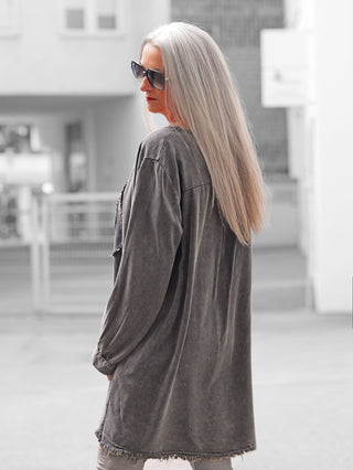 Person in urbanem Umfeld trägt die OFF#DLY Tunica PEACE anthrazit, ein lässiges Longshirt mit fransigem Saum und großem glitzernden Peace-Zeichen, über hellen Hosen.
