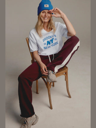 Frau in locker sitzender Penn&Ink Pant aubergine mit weißen Seitenstreifen, elastischem Bund und Kordelzug. Sie sitzt lässig auf einem Stuhl, trägt weiße Sneakers und eine blaue Mütze.