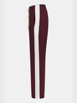 Penn&Ink Pant aubergine: Seitlich fotografierte burgunderfarbene Trackpants mit breitem weißen Streifen, elastischem Bund mit Kordelzug und sichtbarer Seitentasche, leicht zulaufend an den Knöcheln, entspannte Passform.