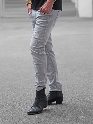 OFF#DLY Denim Sergnano grey Jeans mit Used-Look und Destroyed-Details, getragen mit schwarzen Western-Stiefeletten. Hände lässig in den Taschen, sichtbare goldene Armreifen und Ringe.