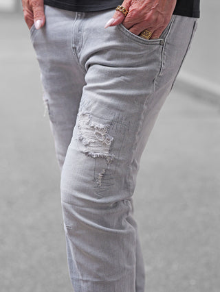 OFF#DLY Denim Sergnano grey: Nahaufnahme von grauen, distressed Jeans mit modischen Rissen am Knie, getragen von einer Person mit goldfarbenem Schmuck. Hände in den Hosentaschen, lässiger urbaner Look.