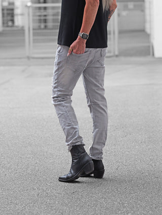 Person trägt OFF#DLY Denim Sergnano grey Jeans, leicht hochgekrempelt, kombiniert mit schwarzen Lederstiefeln. Hand mit goldenen Ringen lässig in der Gesäßtasche, stehend auf gepflastertem Untergrund.