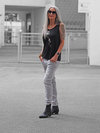 Frau trägt OFF#DLY Denim Sergnano grey: graue, leicht zerrissene Jeans im Used-Look, kombiniert mit einem schwarzen Tanktop und schwarzen Stiefeln, ergänzt durch Accessoires und Tattoo.