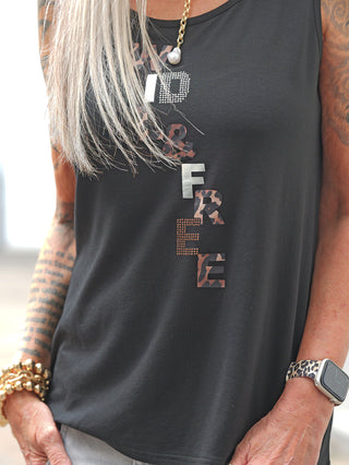Person trägt Doris Streich Top 552130 in Schwarz, mit WILD & FREE in metallischen, getarnten Buchstaben. Sichtbar sind tätowierte Unterarme, goldene Armbänder und eine Kette mit Anhänger.