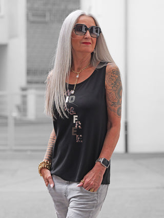 Ältere Frau in einem Doris Streich Top 552130 schwarz mit großem Buchstaben-Print, kombiniert mit Goldschmuck und Tattoos, steht lässig mit Händen in Taschen in urbaner Umgebung.