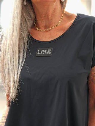 Nahaufnahme des Doris Streich Shirt 513600 schwarz mit kurzen Ärmeln, verziert mit einem LIKE-Patch aus Strasssteinen auf der Brust. Ein goldener Kettenanhänger und eine Perlenohrring runden den Look ab.