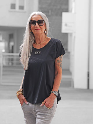 Silberhaarige Frau trägt Doris Streich Shirt 513600 schwarz, locker geschnitten mit kleinem LIKE-Patch, kombiniert mit auffälligem Goldschmuck und einer Smartwatch. Jeans leicht zerstört, urbanes Ambiente im Hintergrund.
