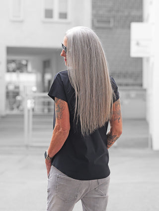 Person von hinten mit langen, silber-grauen Haaren trägt das Doris Streich Shirt 513600 in Schwarz, kombiniert mit hellgrauen Jeans. Sichtbare Tattoos auf den Unterarmen. Hintergrund unscharf.