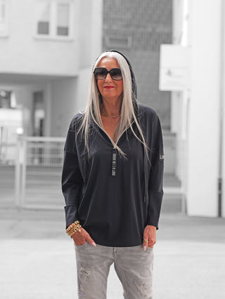 Frau mit langen, silberweißen Haaren trägt eine Doris Streich Tunika 401600 in Schwarz, kombiniert mit goldenen Accessoires und hellgrauen, leicht zerrissenen Jeans, in einer urbanen Umgebung.