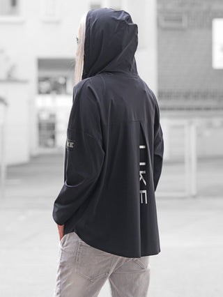 Die Rückansicht zeigt eine Person in einer Doris Streich Tunika 401600 black, einem schwarzen, lockeren Windbreaker mit Kapuze und LIKE Aufdruck, kombiniert mit hellgrauen Jeans.