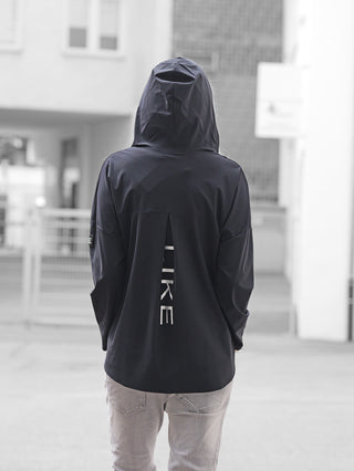 Eine Person trägt eine schwarze Doris Streich Tunika 401600 mit Kapuze und einem vertikalen NIKE-Schriftzug auf dem Rücken, kombiniert mit hellgrauen Jeans, in einer urbanen Umgebung.