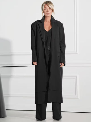 Gitta Banko Mantel Athena black: Frau trägt langen, einreihigen schwarzen Mantel mit Reverskragen, über schwarzem Paillettentop und weiten Hosen. Minimalistischer, schicker Look mit entspannter Pose.