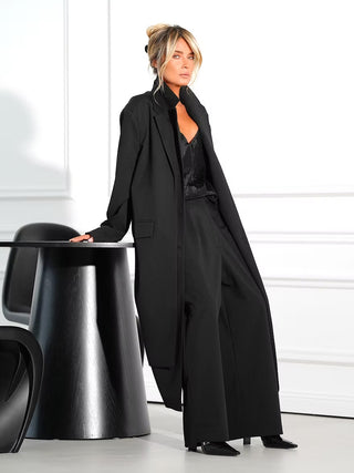 Gitta Banko Mantel Athena black: Frau trägt langen, offenen schwarzen Mantel mit Revers über schwarzer Hose und Top, lehnt an schwarzem Tisch, modernes und elegantes Erscheinungsbild.
