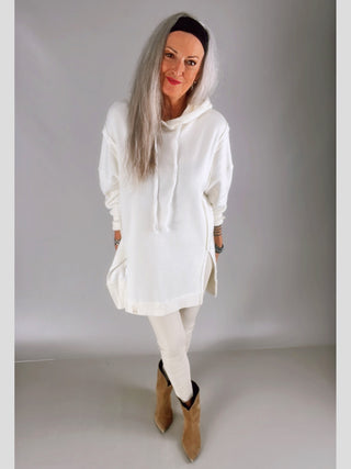Frau trägt Cotton Candy Sweatshirt Luna offwhite, ein übergroßes, langes Hoodie mit Seitenschlitzen und Fronttasche, kombiniert mit passenden Leggings und tanfarbenen Stiefeletten, für einen lässigen Look.