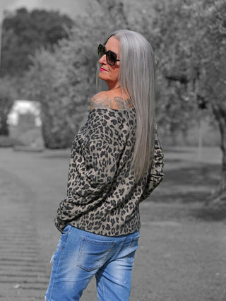 Frau mit langen, silbergrauen Haaren trägt die OFF#DLY Jacke Linnea Leo green black, ein schulterfreier Leo-Print Pullover, der über eine schwarze Jeans drapiert ist. Brillen und Lippenstift ergänzen den Look.