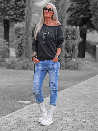 Frau im Park trägt das OFF#DLY Shirt Smile black, ein schwarzes Off-Shoulder-Sweatshirt mit SMILE-Aufdruck, kombiniert mit hellblauen, zerrissenen Jeans und weißen High-Top-Stiefeln.
