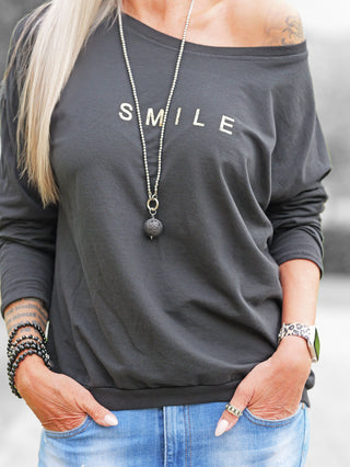 Person trägt das OFF#DLY Shirt Smile black, ein lockeres, schulterfreies Oberteil mit SMILE-Aufdruck. Kombiniert mit langen Perlenkette und Jeans, ergänzt durch Armbänder und Uhr mit Leopardenmuster.