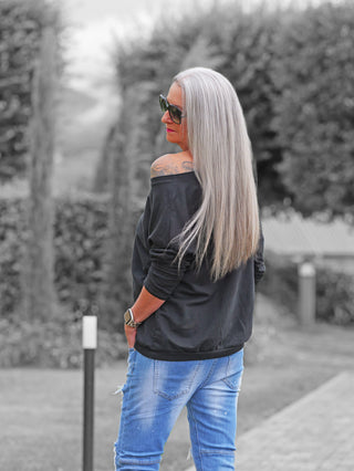 Person mit langen, silbergrauen Haaren trägt ein OFF#DLY Shirt Smile black, ein schwarzes, off-shoulder Langarmshirt, kombiniert mit einer hellblauen Jeans und einer rechteckigen Smartwatch am Handgelenk.