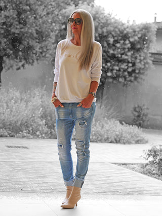 Erwachsener mit dem OFF#DLY Shirt Smile white: weißer Pullover mit SMILE-Aufdruck, kombiniert mit distressed Jeans, beigen Stiefeletten und Accessoires, auf einem nassen, gepflasterten Hof stehend.