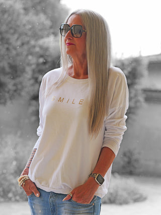 Person mit langen grauen Haaren trägt das OFF#DLY Shirt Smile weiß mit SMILE in Metallic-Schrift. Kombiniert mit einer Apple Watch und lässigen Jeans, vermittelt es entspannte Vibes.