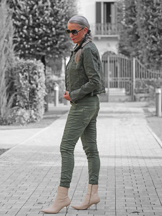 Eine schlanke Frau trägt die OFF#DLY Color Jeans Imelda khaki, kombiniert mit einer olivgrünen Jacke und beige Stiefeletten, während sie auf einem Gartenweg steht.