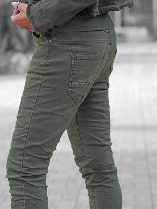 Person trägt OFF#DLY Color Jeans Imelda khaki, schmal geschnitten mit sichtbarer Gesäßtasche. Linke Hand mit Ringen am Oberschenkel. Kombiniert mit passender Jacke für einen urbanen Look.