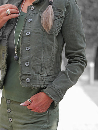 Nahaufnahme eines Torsos mit der OFF#DLY Jacke Bologna verde, einem olivgrünen, fransigen Denim mit Taschen und Knopfleiste, kombiniert mit passenden Ringen und einer langen Kettenhalskette.