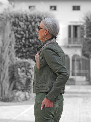 Ältere Frau im Profil trägt die OFF#DLY Jacke Bologna verde aus olivgrünem Denim mit Knopfleiste und ausgefransten Säumen, kombiniert mit passender Hose, Hand in der Tasche.