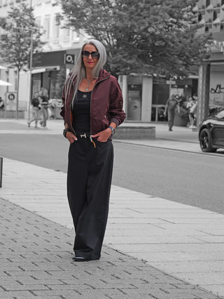 Die Gitta Banko Hose Taki in schwarz präsentiert sich mit einer eleganten Wide-Leg-Passform und hoher Taille, getragen von einer Frau mit burgunderfarbener Bomberjacke auf einer Stadtstraße.