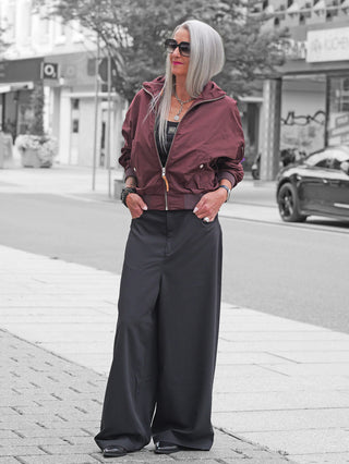 Frau in Gitta Banko Hose Taki schwarz mit weitem Schnitt und hoher Taille, kombiniert mit burgunderroter Jacke, breiten schwarzen Sonnenbrillen und silbernen Accessoires auf städtischem Gehweg.