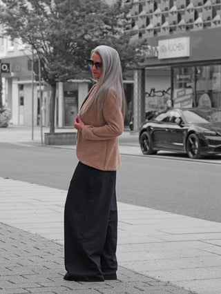 Frau in weiter schwarzer Gitta Banko Hose Taki mit eleganter Silhouette und hoher Taille, kombiniert mit einem camel-farbenen Blazer, auf einem städtischen Gehweg stehend.