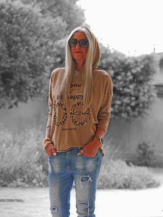 Person im Garten trägt das OFF#DLY Shirt Be Happy camel: ein locker sitzender, tan-brauner Hoodie mit happy und RebelQueen Print, kombiniert mit hellblauen, zerrissenen Jeans.