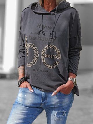 Person trägt den OFF#DLY Shirt Be Happy anthrazit Hoodie mit Be-Leopardenprint und REBELQUEEN-Aufschrift, kombiniert mit mittelblauen Jeans. Ergänzt wird der Look durch dunkle Armbänder und eine Smartwatch.