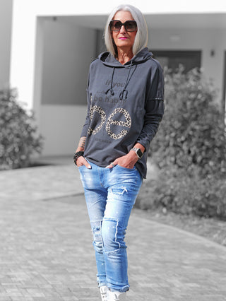 Frau trägt OFF#DLY Shirt Be Happy anthrazit, ein locker sitzendes, graues Hoodie mit „if you want be happy“ und Leoparden-Print, kombiniert mit hellen Jeans und weißen Turnschuhen.
