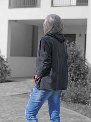 Person von hinten mit schulterlangem grauem Haar trägt das OFF#DLY Shirt Be Happy anthrazit, ein lockeres, dunkles Hoodie mit Kapuze und lässigem Schnitt, kombiniert mit schmalen, leicht zerrissenen Jeans.