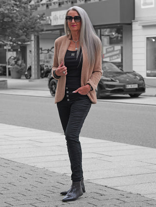 Schlanke Frau trägt OFF#DLY Color Jeans Imelda black, Slim-Fit mit Knopfdetails. Kombiniert mit schwarzem Top und Stiefeletten, akzentuiert durch Accessoires. Urbaner Chic auf einer Stadtstraße.