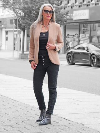 Frau in OFF#DLY Color Jeans Imelda black, Slim-Fit, mit sichtbaren Knöpfen, kombiniert mit camel Blazer und schwarzen Stiefeletten, präsentiert moderne, komfortable Denim-Optik.