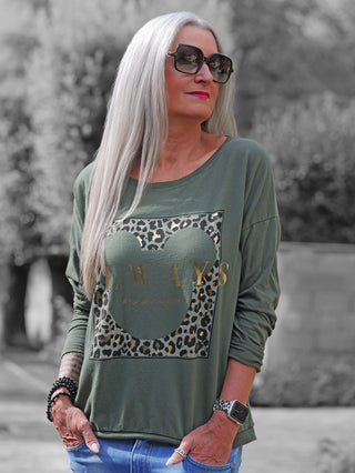 Person mit langen, silbernen Haaren trägt das OFF#DLY Longsleeve Always khaki mit Mickey-Ohren-Print und Leopardenmuster, kombiniert mit blauen Jeans und Armbanduhr.