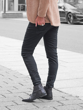 Person trägt OFF#DLY Color Jeans Imelda black, schlanke Passform, betont Figur, kombiniert mit schwarzen Lederstiefeln und einem camel-farbenen Pullover, Hand mit silbernem Ring sichtbar.