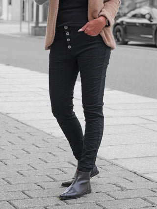 OFF#DLY Color Jeans Imelda black in Slim-Fit mit sichtbarem Vier-Knopf-Verschluss, kombiniert mit dunklen Lederstiefeln und beigefarbener Strickjacke, getragen auf einem Gehweg neben einer Straße.