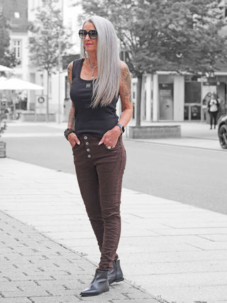 Frau auf Bürgersteig trägt OFF#DLY Color Jeans Imelda schoko, eine figurbetonte, hochgeschnittene Hose mit Knopfleiste, kombiniert mit schwarzem Tanktop und Stiefeletten, betont durch auffällige Accessoires.